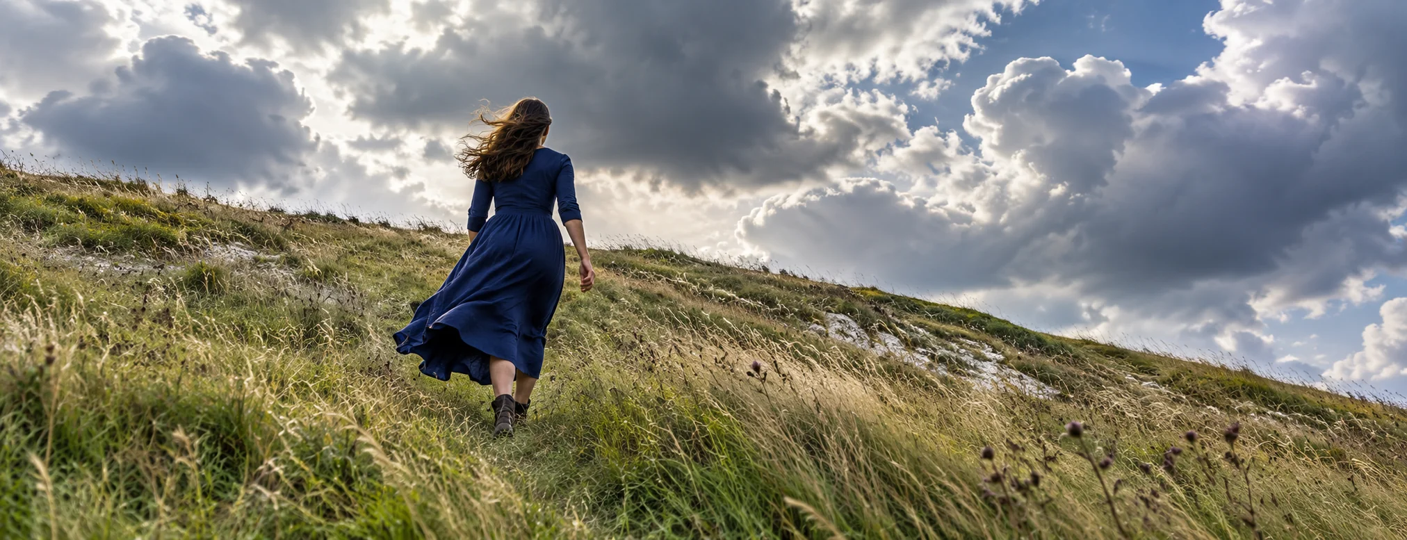 A woman moves up a grassy chalk down beneath clouds